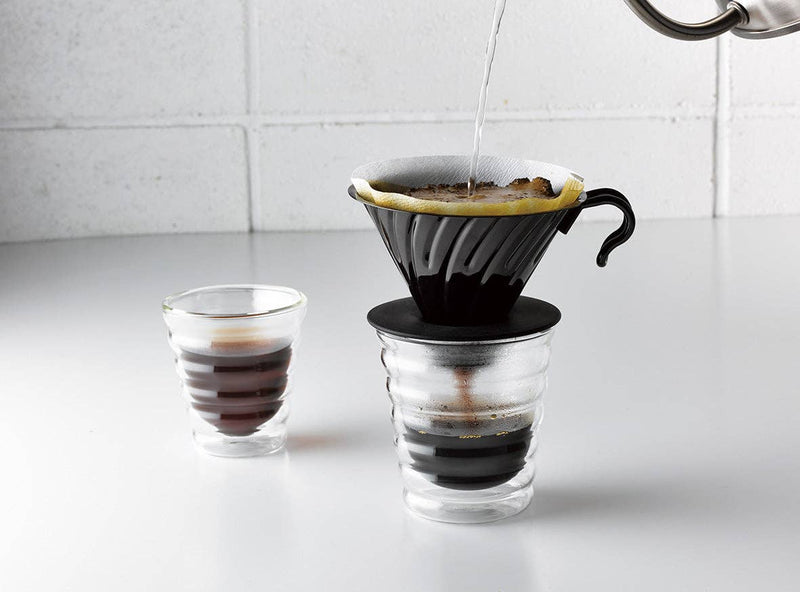 HARIO USA - V60 Metal Dripper: Silver in use, brewing coffee into a double-walled glass with hot water pouring from a kettle above.