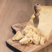 Nicola's Marketplace - Parmigiano Reggiano DOP Mitica® 36 months: 7 Ounces cheese on a wooden board with knife, showcasing its granular texture and crumbly chunks.