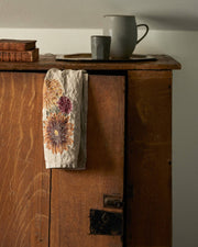 Coral & Tusk - Autumn Sunflower Tea Towel draped on rustic cabinet, featuring embroidered autumnal blooms; embodies cozy, seasonal warmth in a kitchen setting.