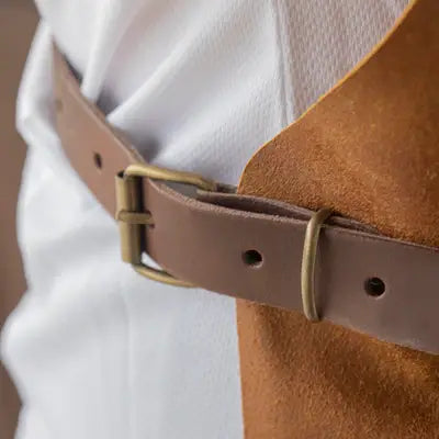 Messermeister - Vintage Leather Apron - Amber, featuring a medium-brown leather belt with brass buckle, styled over a white shirt and camel suede layer.