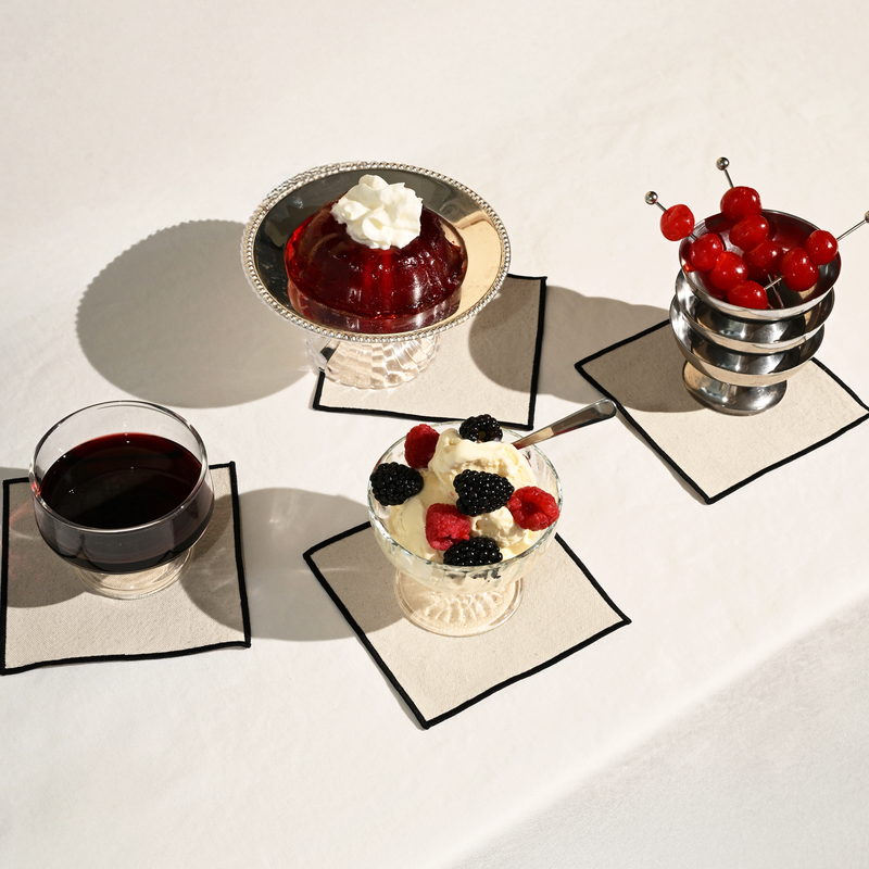 ATELIER SAUCIER - Cream Burlap Noir Cocktail Napkins: Displayed with desserts, showcasing a set of four durable, cream burlap napkins with black edging on a tabletop.