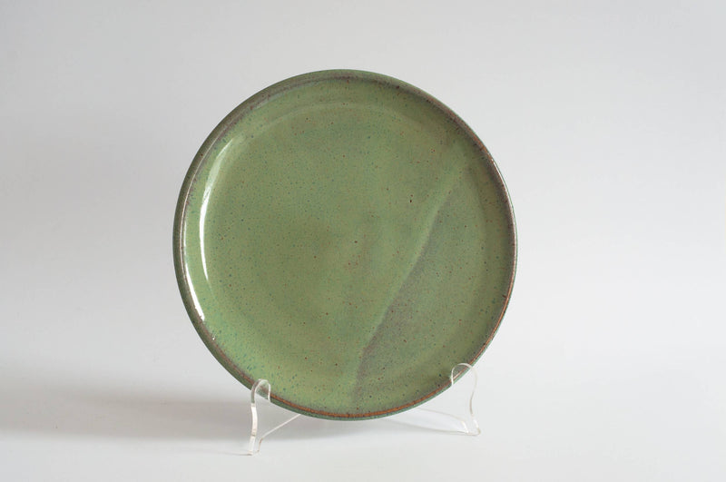 RachaelPots - Dinner Plate: Coral, a round ceramic plate with a muted green speckled glaze, stands upright, showcasing its handmade craftsmanship and slightly upturned lip.