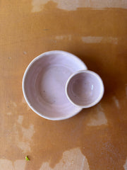 Top-down view of ROB Ceramics' handmade olive serving bowl in dusty-pink, featuring a small attached bowl for depositing pits, on a rustic tabletop.