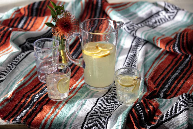 Zwiesel Fortessa Tritan Outdoor Pitcher in clear polycarbonate, filled with lemonade and lemon slices, displayed on a picnic setup with glasses and a flower vase.