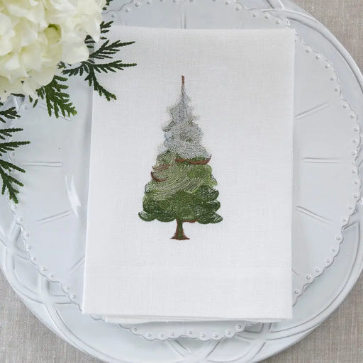 Tri-Fold Snowy Pine Napkin by Arte Italica & Crown Linen Designs, featuring an embroidered evergreen motif, elegantly folded on a decorative plate with cedar sprig and hydrangea accents.