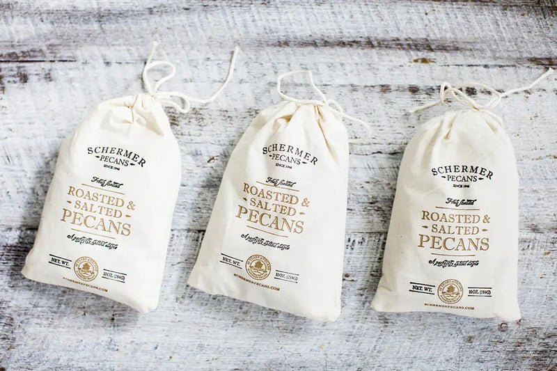 Schermer Pecans - ROASTED AND SALTED PECANS - Ga Grown - 12 cloth bags displayed on a wooden surface, emphasizing fresh, hand-selected pecans from Georgia orchards.