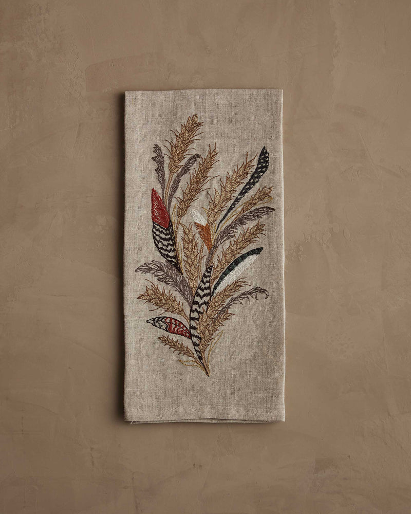 Coral & Tusk - Wheat Harvest Tea Towel featuring embroidered autumnal grasses and feathers, folded on a tan surface. Perfect for seasonal kitchen decor.