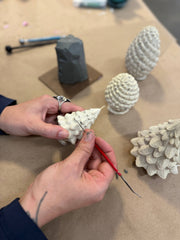 Hands sculpting Mo&Co Home exclusive beeswax tree candle, inspired by vintage ornaments. Table holds more sculptures, clay, and tools, embodying meticulous craftsmanship.