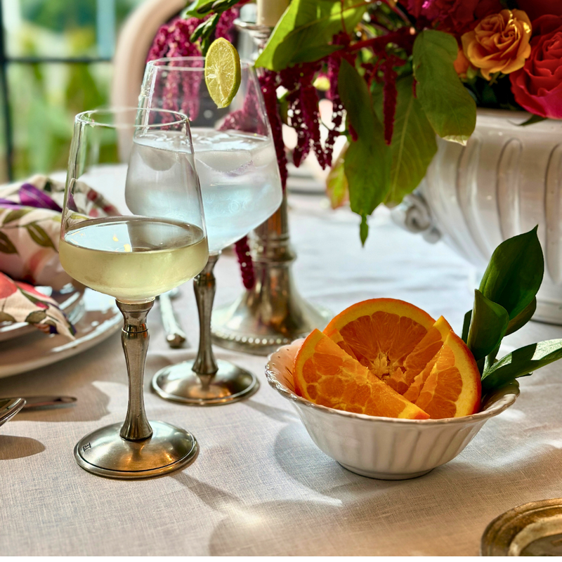 Arte Italica & Crown Linen Designs Bella Bianca Beaded Cereal Bowl on a table with wine glasses and sliced fruit, showcasing elegant Italian craftsmanship.