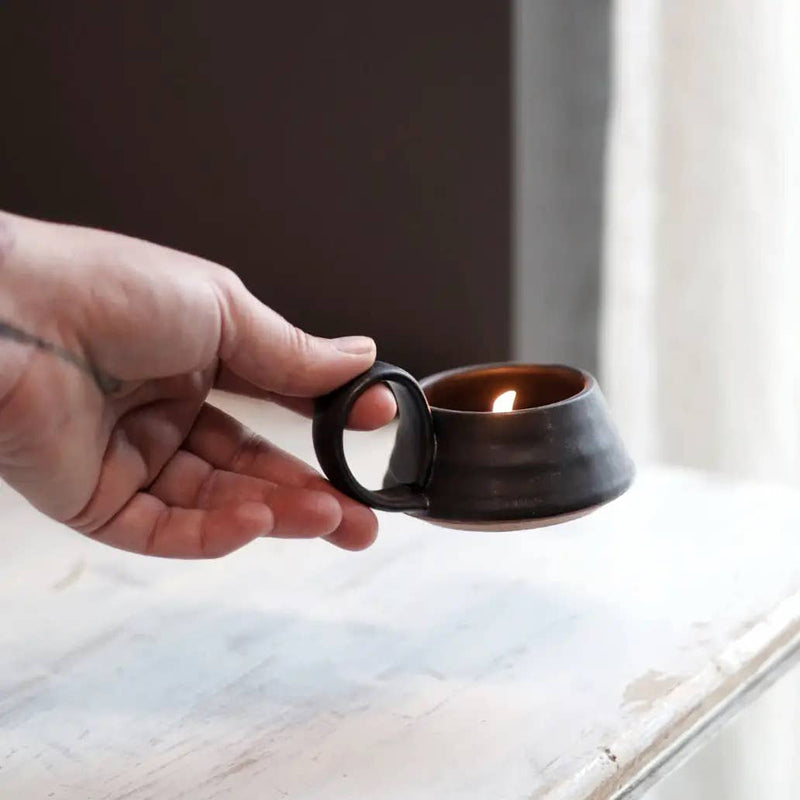 Hand holding Gravesco Pottery - Tea Lite Candle Holder in Black, featuring a flickering candle flame, showcasing its portable, wheel-thrown stoneware design with a classic loop handle.