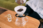 HARIO USA - HARIO V60 Glass Dripper 02 with Olive Wood holder, shown brewing coffee in a pour-over setup with a glass carafe on a wooden table.