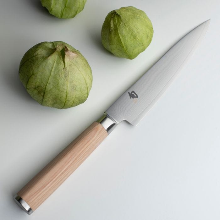 Shun Classic Blonde Utility Knife, 6 inches, with a Damascus-pattern blade and wooden handle, lies beside green tomatillos on a white surface.