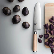 Shun Classic Blonde Santoku, 7 on a countertop, with wooden handle and Damascus-pattern blade, placed between whole and chopped purple potatoes on a cutting board.