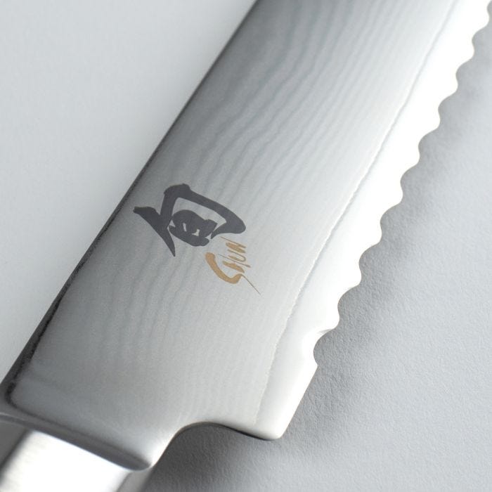Close-up of Shun Classic Bread Ebony knife blade, showcasing Damascus-style waves, scalloped serrations for effective bread slicing, and a black and gold maker’s mark.