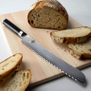 Shun | Classic Bread Ebony, 9 inches knife, with scalloped edge, rests on a cutting board beside a sliced crusty loaf, highlighting precise slicing capability.