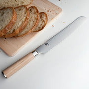 Shun Classic Blonde Bread Knife, 9 inches, with a light wood handle, lies beside a cutting board with sliced crusty bread and scattered crumbs.