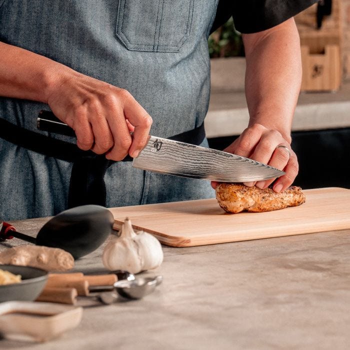 Shun Classic Chef's Ebony Knife, 8-inch, expertly slices chicken on a wooden board in a kitchen scene, highlighting its versatile, ergonomic design and superior cutting performance.