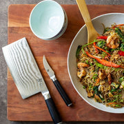 Shun Classic Vegetable Cleaver, 7 inches, on a cutting board with a nakiri knife, bowls, and a serving bowl of stir-fried vermicelli with vegetables and shrimp.