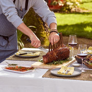 Person carving roast with Shun Classic 2-Piece Carving Set featuring a slicing knife and fork, amidst an elegant outdoor dining setting with wine and side dishes.