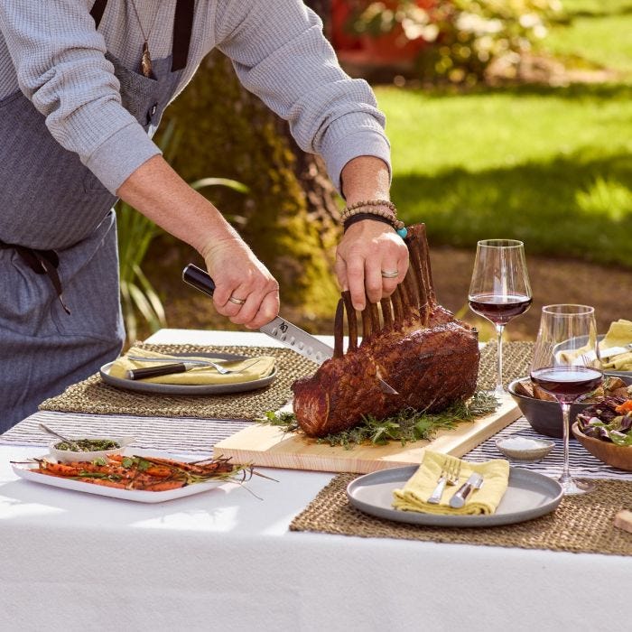 Person carving roast with Shun Classic 2-Piece Carving Set featuring a slicing knife and fork, amidst an elegant outdoor dining setting with wine and side dishes.