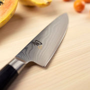 Shun Classic Chef's Ebony, 6-inch knife with Damascus-style blade on wooden board, near halved papaya and small orange fruits, embodying precision and versatility.