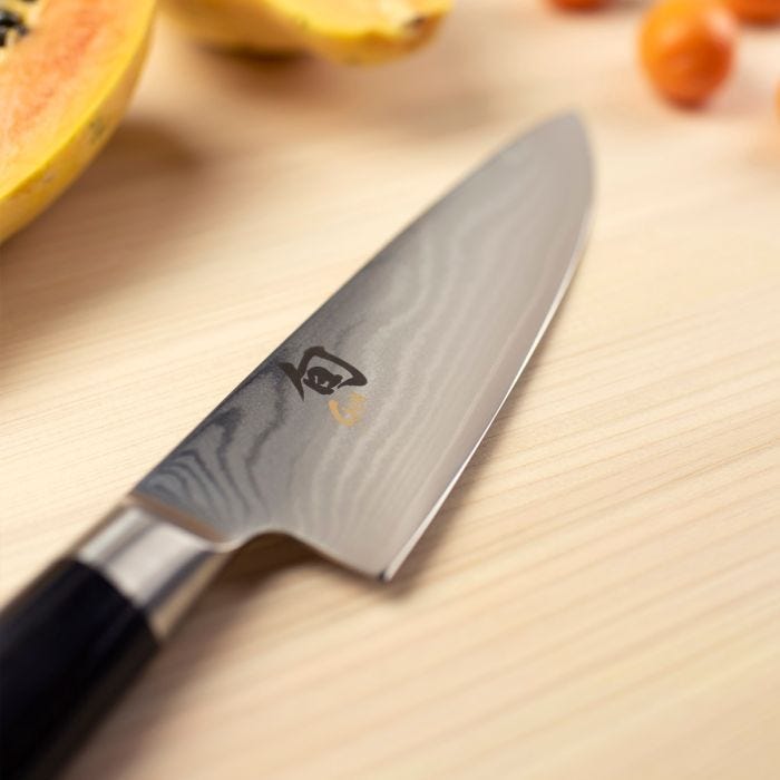 Shun Classic Chef's Ebony, 6-inch knife with Damascus-style blade on wooden board, near halved papaya and small orange fruits, embodying precision and versatility.