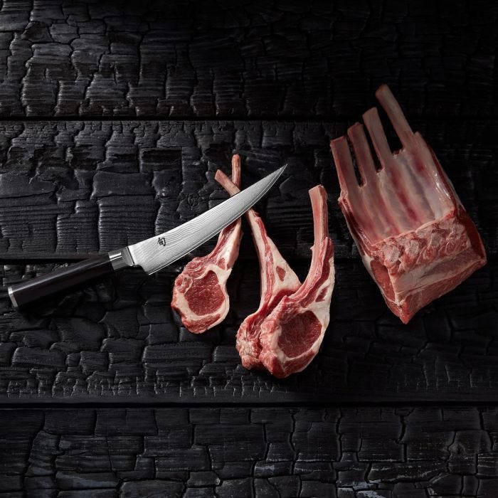 Shun | Classic Boning & Fillet knife, 6 inches, lies beside raw lamb rib chops on charred wood, showcasing its precision cutting capability for meats.