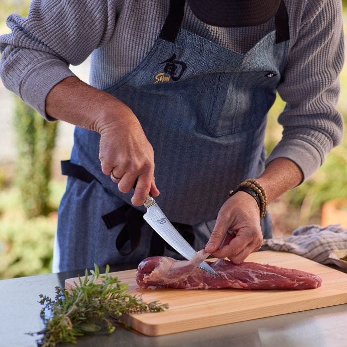 Person in apron trims meat with Shun | Classic Boning & Fillet, 6 inches knife on wooden board; herbs nearby.