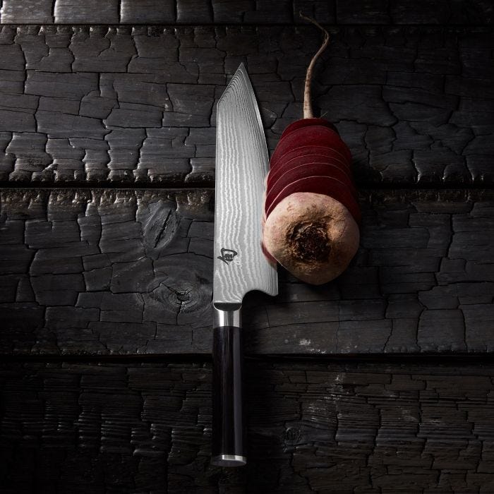 Shun Classic Kiritsuke 8-inch knife with Damascus pattern and dark handle on wood surface, slicing thin beet rounds, showcasing precision and versatility for expert chefs.