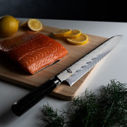 Shun Dual Core Yanagiba knife with Damascus-pattern blade and wooden handle, displayed on a cutting board with salmon, lemon slices, and dill, showcasing elegance and precision.