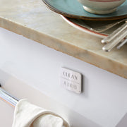 Sir|Madam Nickel-Plated Clean/Dirty Dishwasher Magnet on a dishwasher, indicating status. Plates, bowls, and flatware on counter, towel on handle.