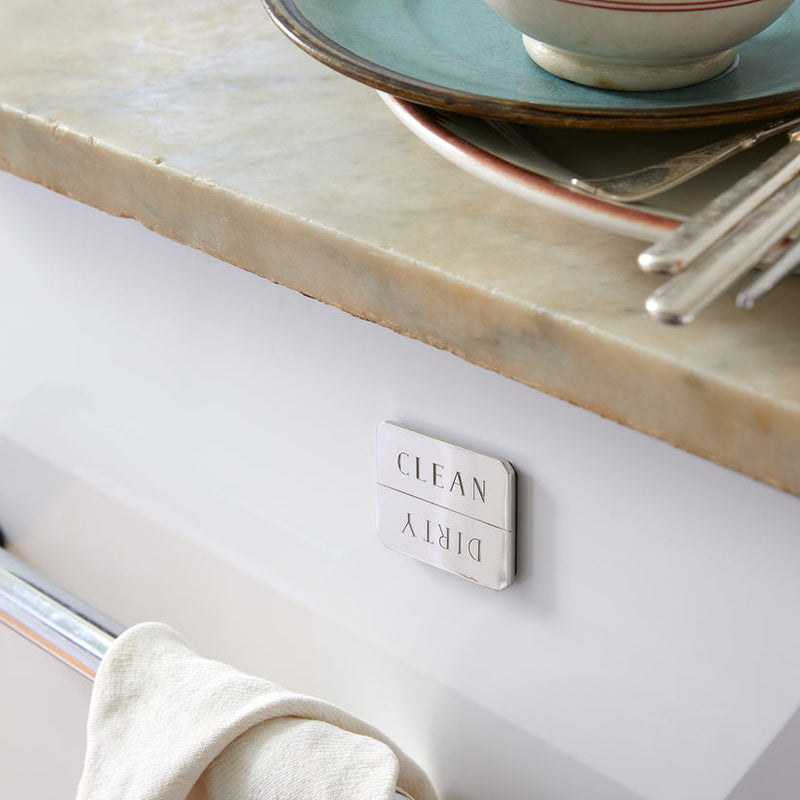 Sir|Madam Nickel-Plated Clean/Dirty Dishwasher Magnet on a dishwasher, indicating status. Plates, bowls, and flatware on counter, towel on handle.