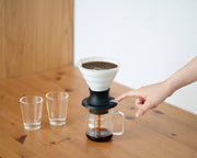 HARIO Ceramic Immersion Dripper 02 SWITCH on table, with a hand adjusting the flow lever. Glass carafe below partially filled with coffee, surrounded by empty glasses.