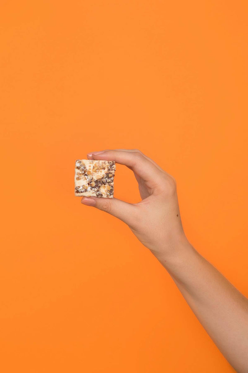 Hand holding a Malvi Mallow - Fluffy Nutter marshmallow square, drizzled with white confection and topped with chopped nuts, against an orange background.