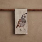 Coral & Tusk - Quail Tea Towel, featuring detailed quail embroidery on natural linen, draped over a wooden rod, ideal for sophisticated kitchen decor.