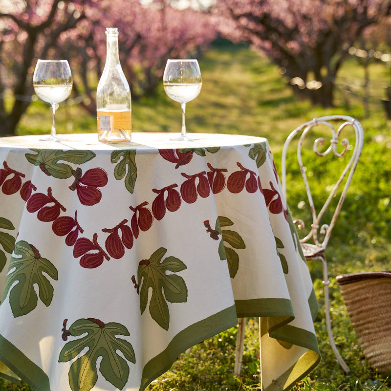 Couleur Nature - Fig Red & Green tablecloth on a round table with wine, enhancing an outdoor picnic ambiance with a bistro chair and basket nearby.