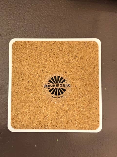 Drinks on Me coaster with cork insert and logo, ideal for adding humor to home bar decor. Perfect for gifting and protecting surfaces. Made in the USA.