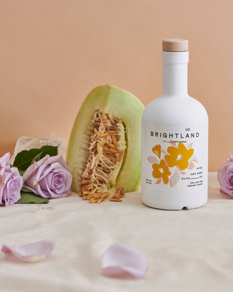 Brightland - ARISE Basil Infused Olive Oil bottle next to melon and flowers, showcasing its artistic label inspired by spring blooms.