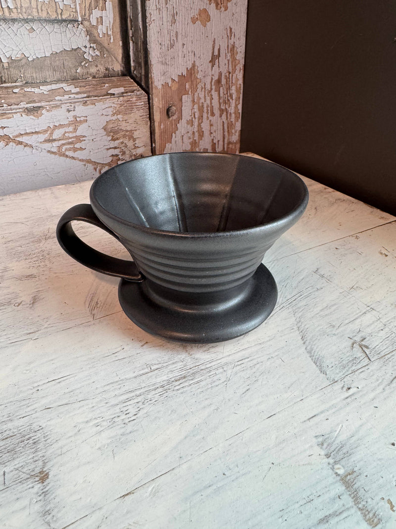 Gravesco Pottery - Handmade Ceramic V60 Coffee Dripper in Black on a rustic surface, showcasing its ridged interior and handle for safe, effective brewing.