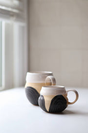Two unique Gravesco Pottery Transformation Mugs, showcasing their handcrafted design, with contrasting glazes, sit on a light surface near a softly lit window.