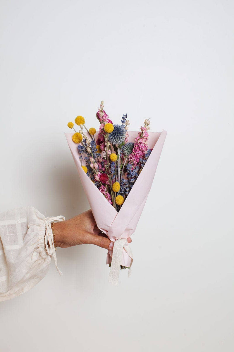 Idlewild Floral Co. - Bright Small Field Bouquet: A hand holds a dried flower bouquet with yellow, blue, and pink blooms, wrapped in pink paper, perfect for a small vase.