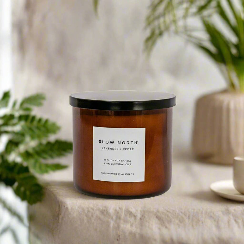 Slow North - Lavender + Cedar - Signature 3-Wick Non-Toxic Candle in amber jar, on cloth with fern and potted plant, ideal for serene, aromatic spaces.