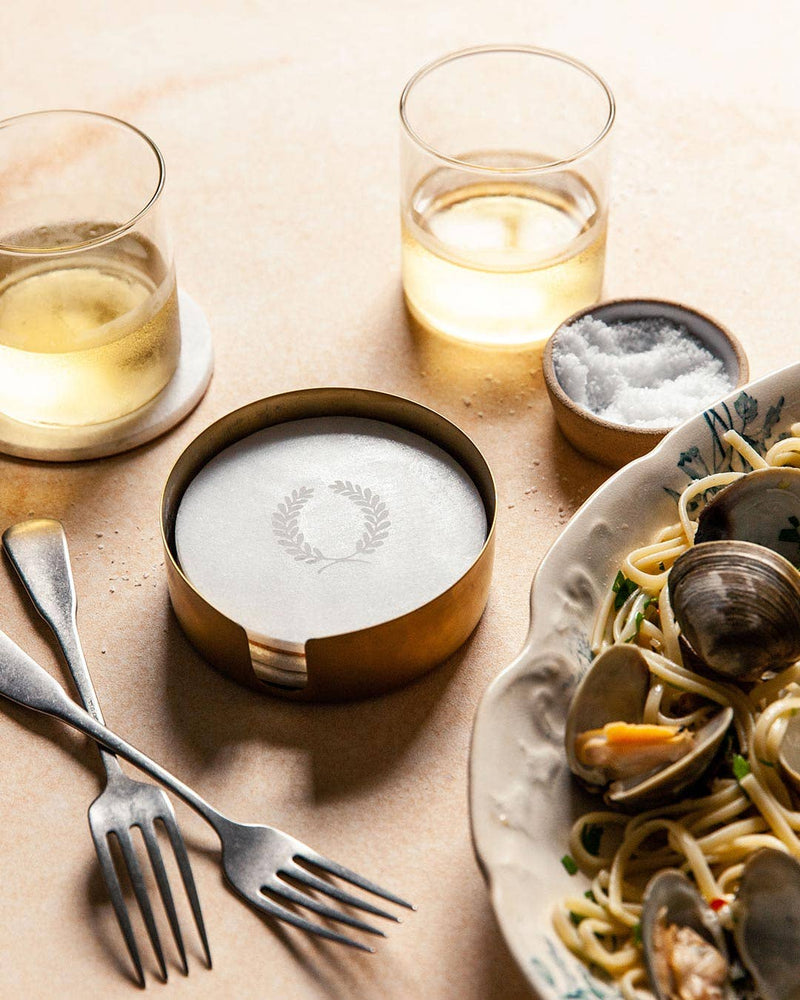 Farmhouse Pottery - Marble Laurel Coasters in a brass holder beside forks and tumblers, set for a seafood pasta meal, embodying Athens Cooks' curated kitchen elegance.