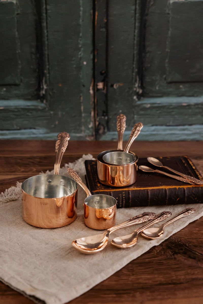 Coppermill Kitchen Vintage Inspired Measuring Cups with ornate handles, displayed on a rustic wooden surface, evoke antique elegance and artisanal craftsmanship.