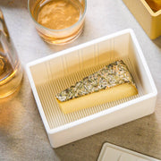 CapaBunga® - Cheese Vault® in white, showcasing a wedge of herb-crusted cheese. A glass of amber-pink liquid is partially visible beside the tray.