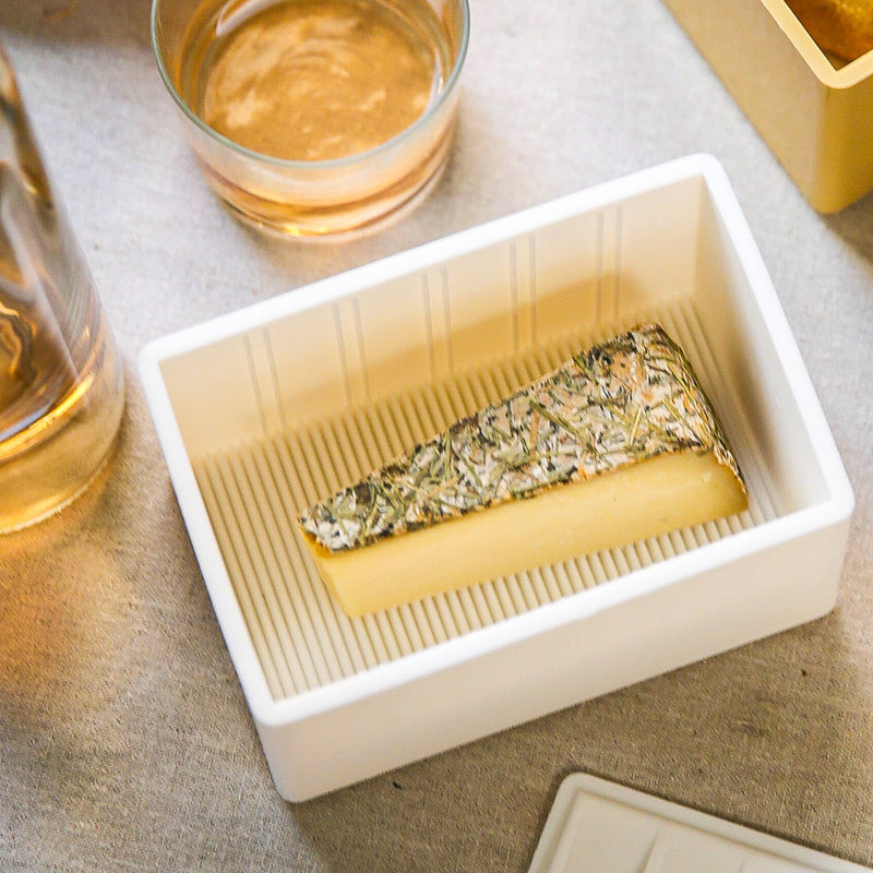 CapaBunga® - Cheese Vault® in white, showcasing a wedge of herb-crusted cheese. A glass of amber-pink liquid is partially visible beside the tray.