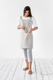Woman wearing MagicLinen's pinafore cross-back linen apron with twin-sectioned front pocket, ideal for kitchen or garden use, available in various colors.