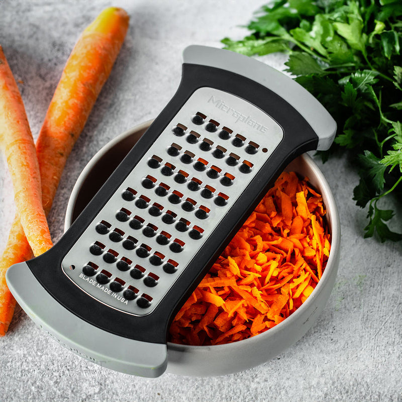 Microplane Bowl Grater resting over a bowl of grated carrot, with whole carrots and herbs nearby; ideal for precise, versatile kitchen prep.