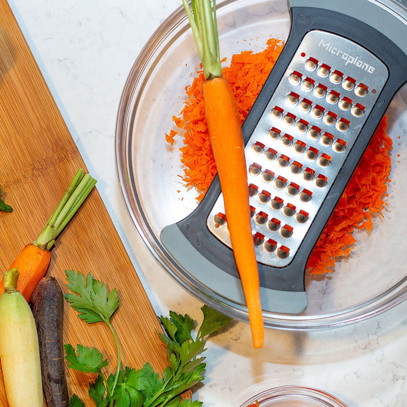 Microplane Bowl Graters, Extra Coarse, showcased grating carrots over a glass bowl, surrounded by colorful carrots and parsley, ideal for efficient meal prep.