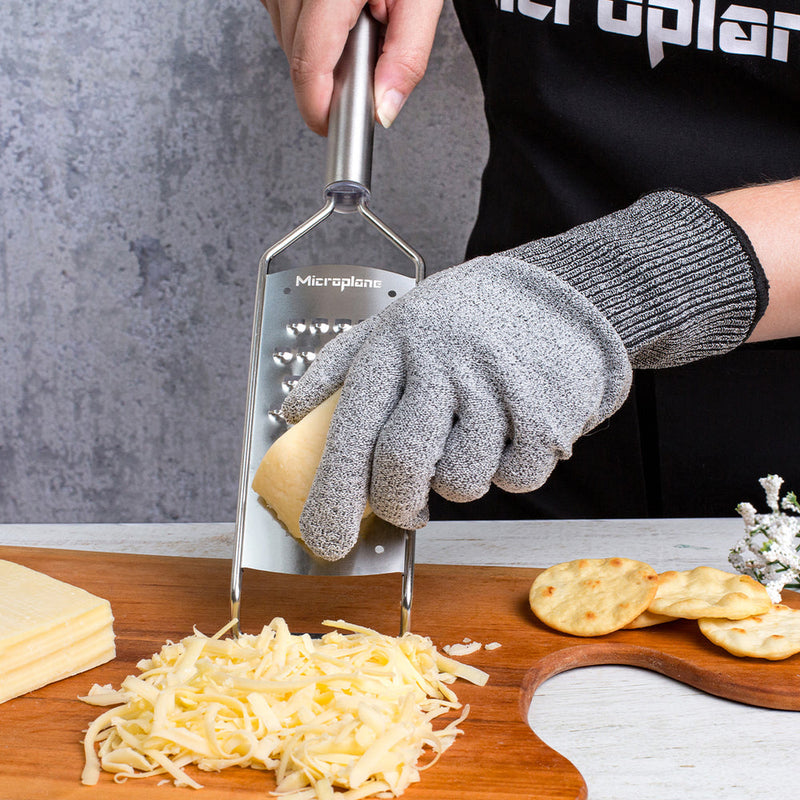 Person uses Microplane | Professional Series Extra Coarse Cheese Grater with cut-resistant glove, shredding cheese onto a cutting board with slices and crackers nearby.
